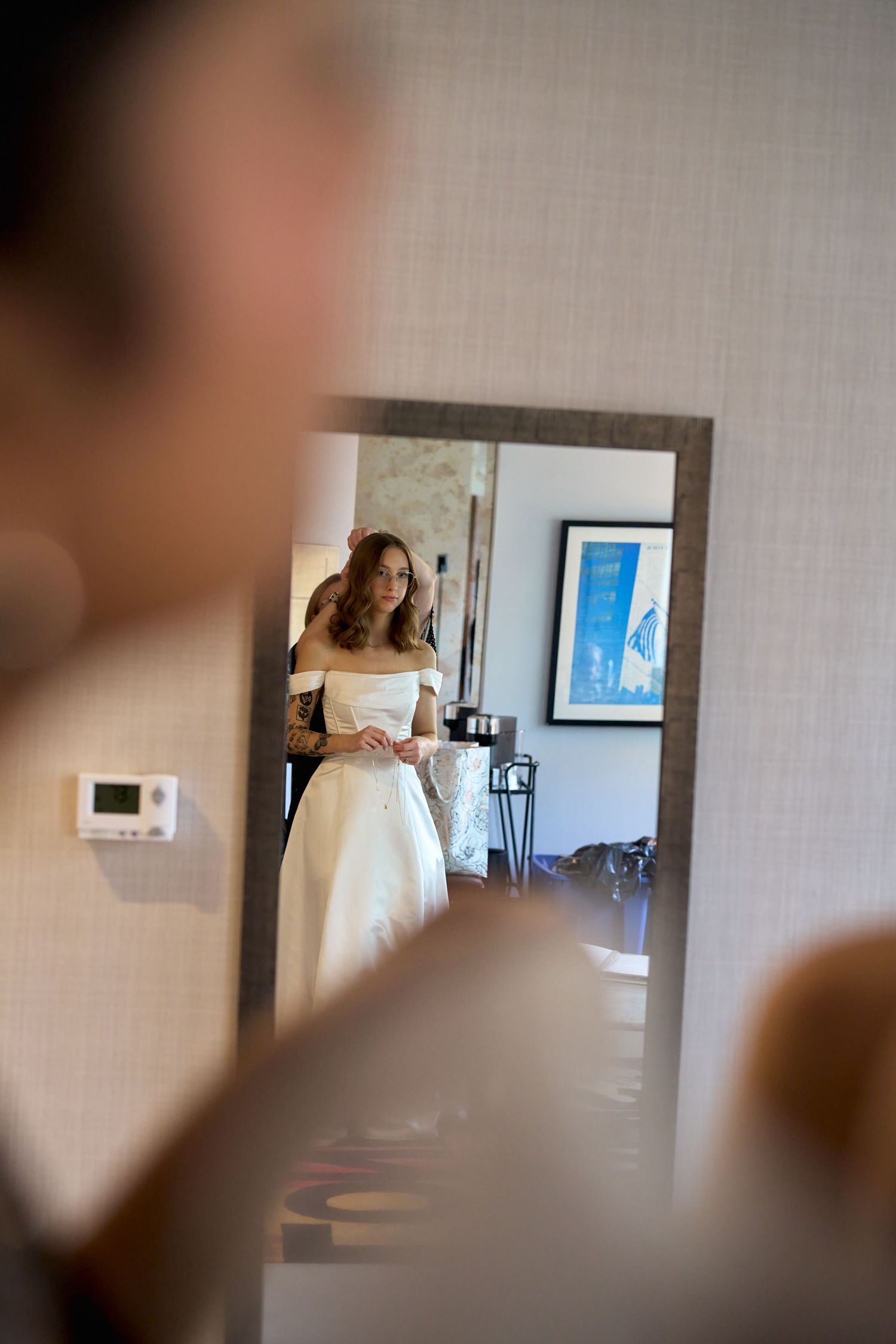 Bride in white wedding dress viewed through mirror reflection in elegant hotel room during bridal preparation.