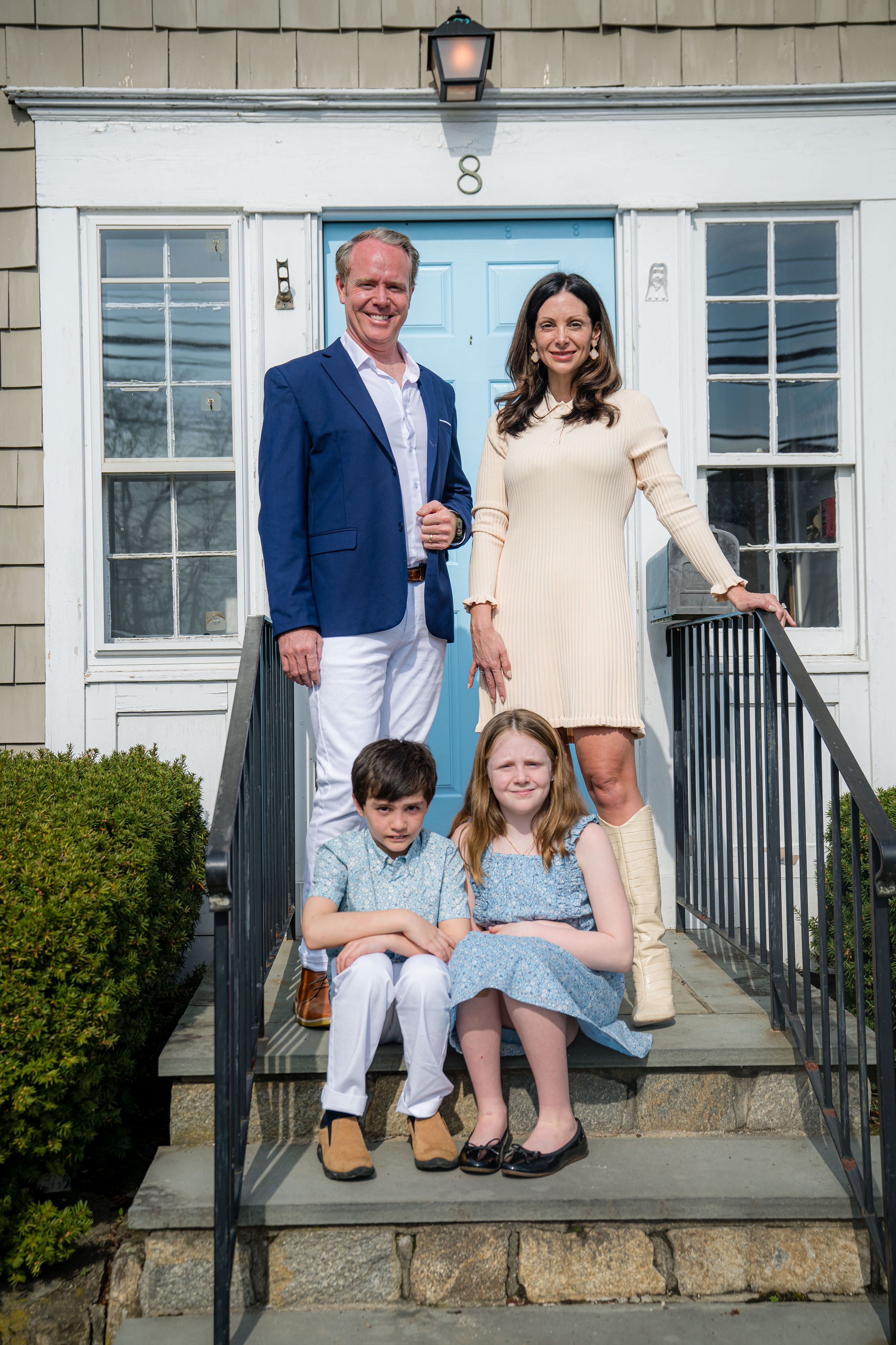Family of four posing on front steps of home with light blue door, parents standing, children sitting on steps.