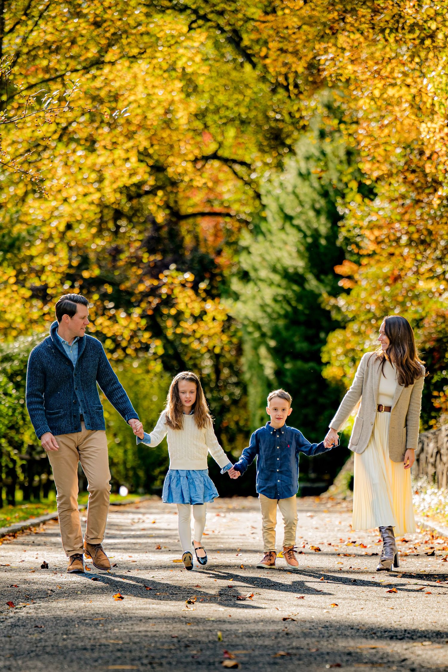 Family of four holding hands walking on a path surrounded by vibrant autumn foliage in a park.