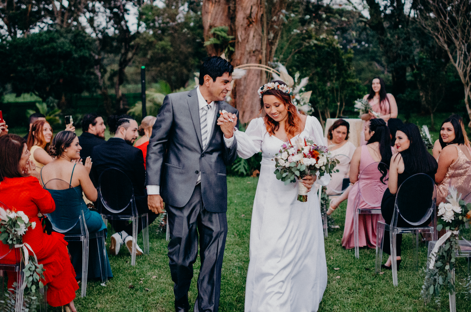 Joyful newlyweds walking down aisle amid outdoor wedding celebration with seated guests.