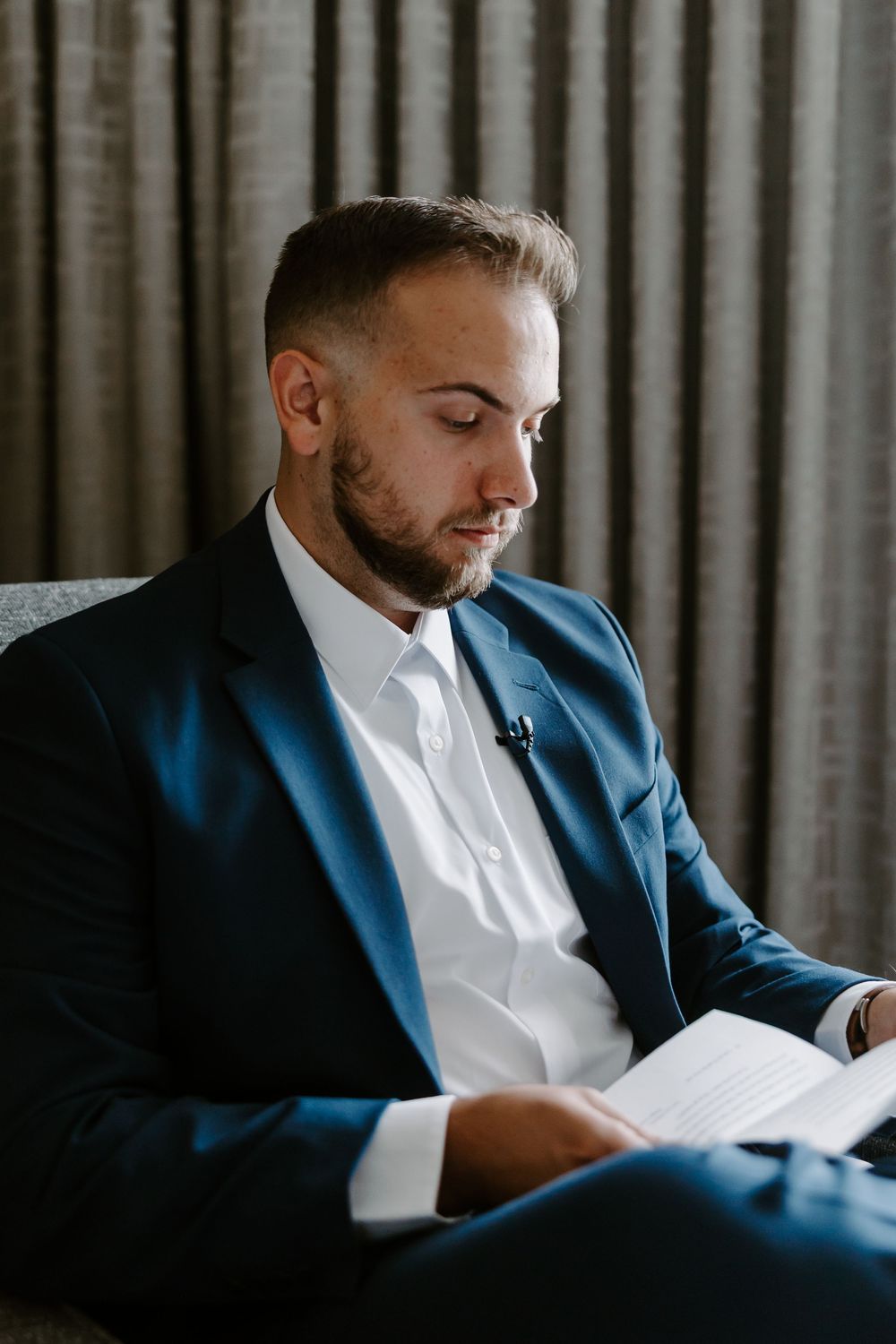 Handsome groom in navy suit reading a heartfelt letter while seated before his wedding ceremony.