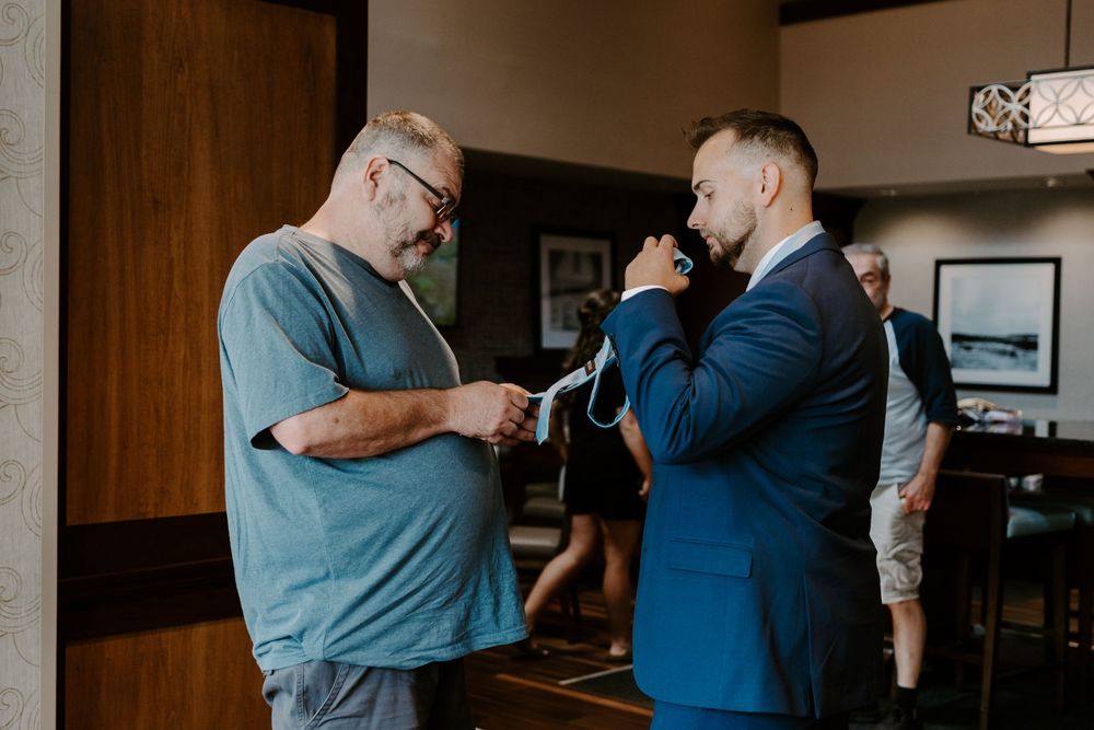 Older gentleman helping a dapper groom adjust his tie during wedding day preparations.