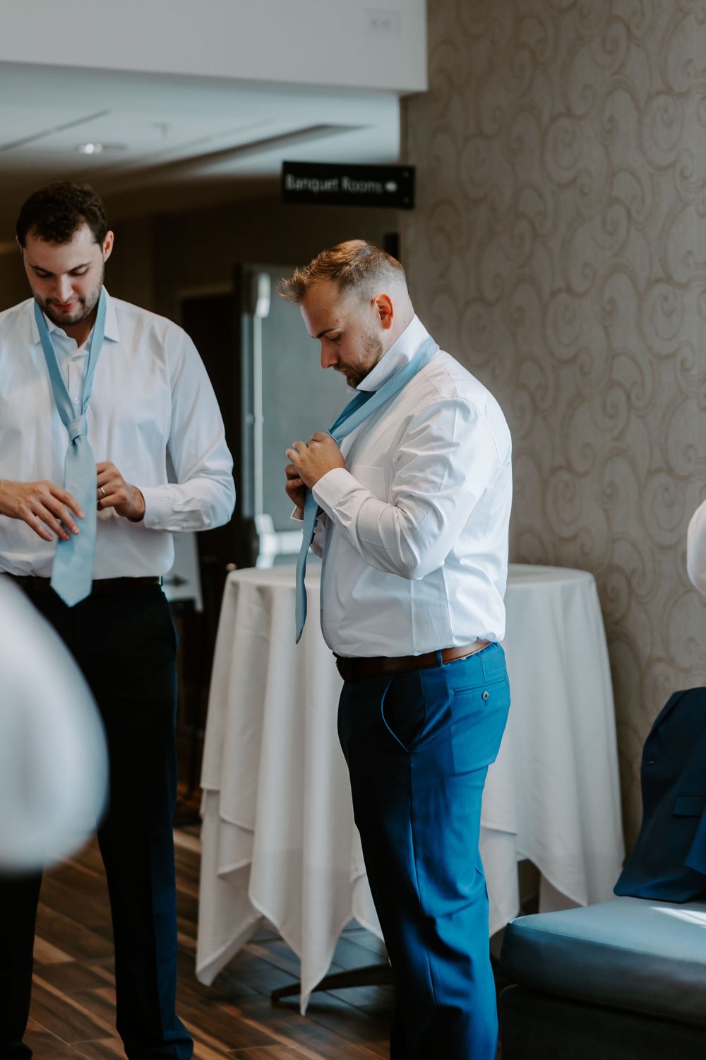 Groomsmen in white shirts and blue ties helping each other get dressed before the wedding.