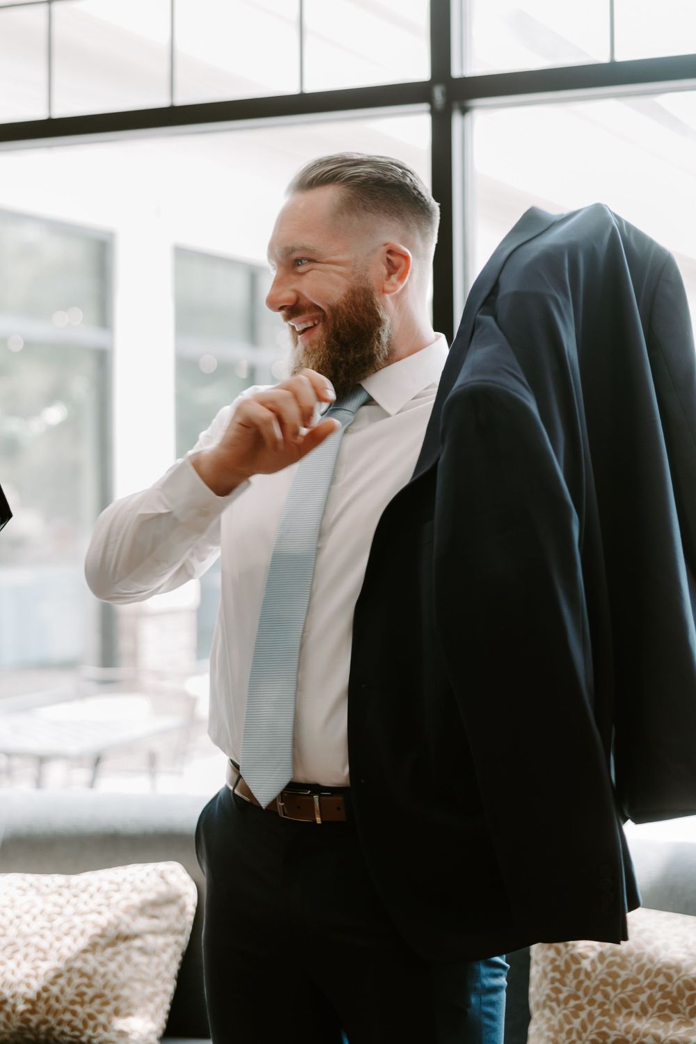 Smiling bearded groom in white shirt and light blue tie preparing his navy wedding suit.