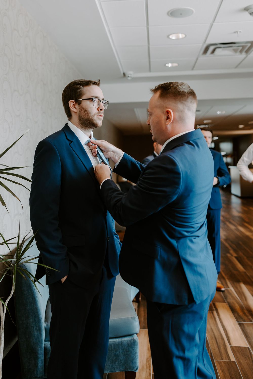 Groomsman in navy suit helping adjust another groomsman's tie before the wedding ceremony.