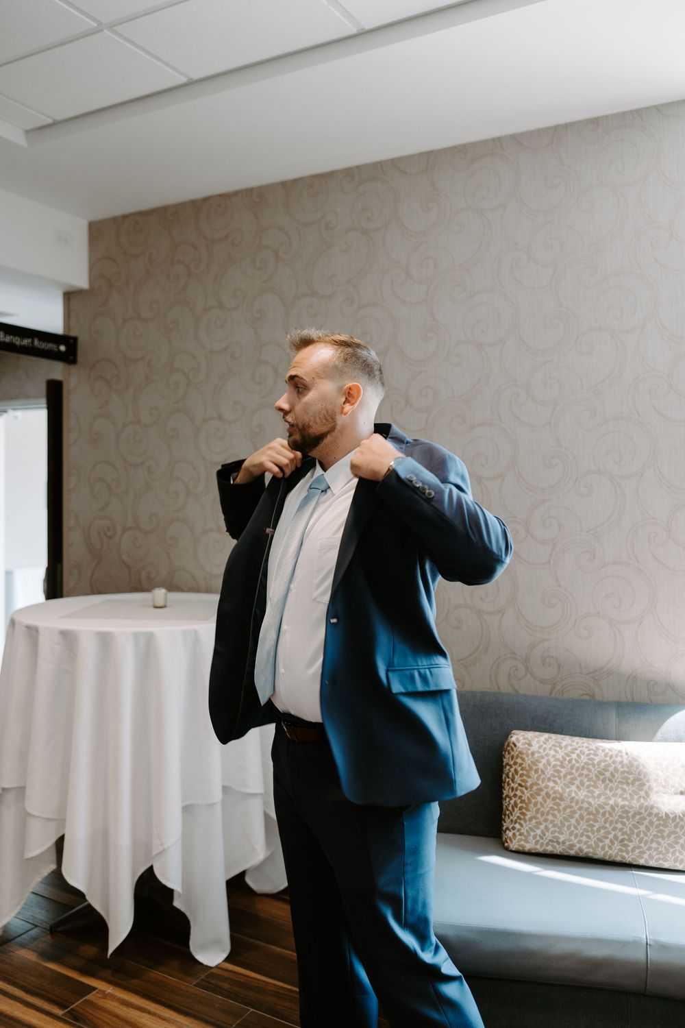 Groom in navy suit adjusting his collar while getting ready in a hotel suite before his wedding.