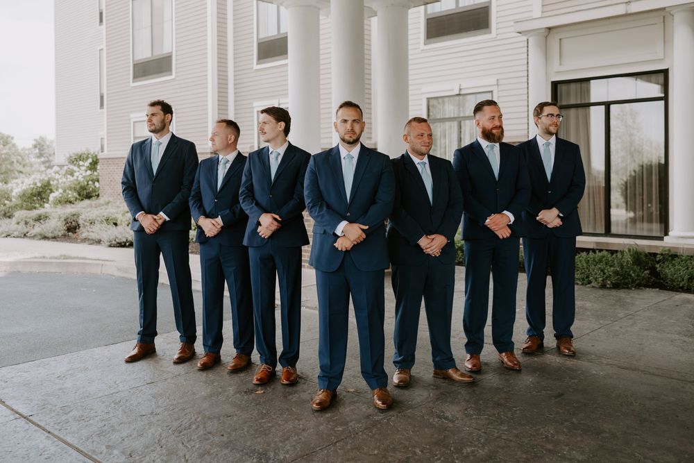 Seven groomsmen in matching navy blue suits with brown shoes posed in a row outside a white building.