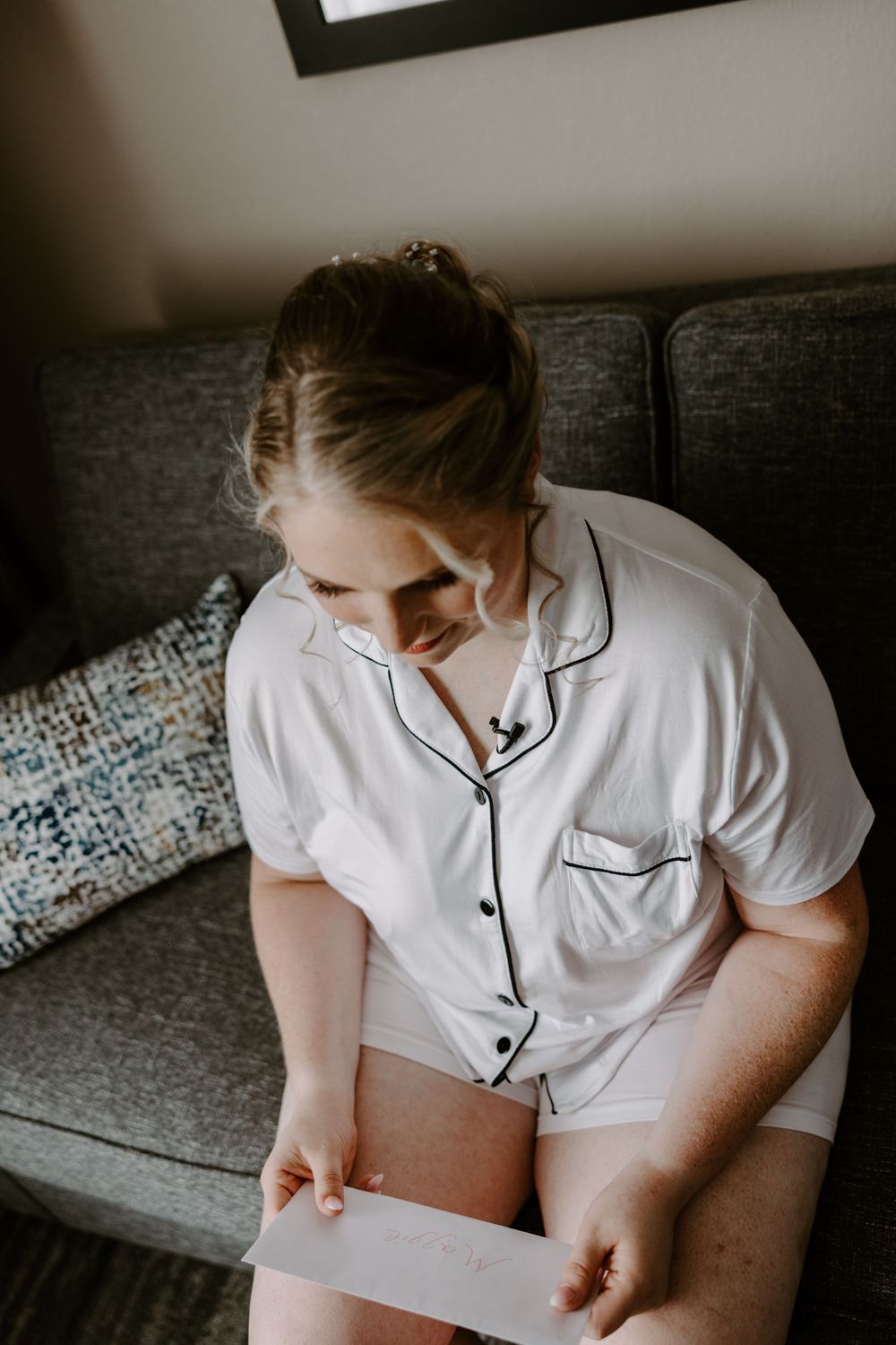 Bride in white pajamas reading a heartfelt letter while seated on a gray couch before her wedding.
