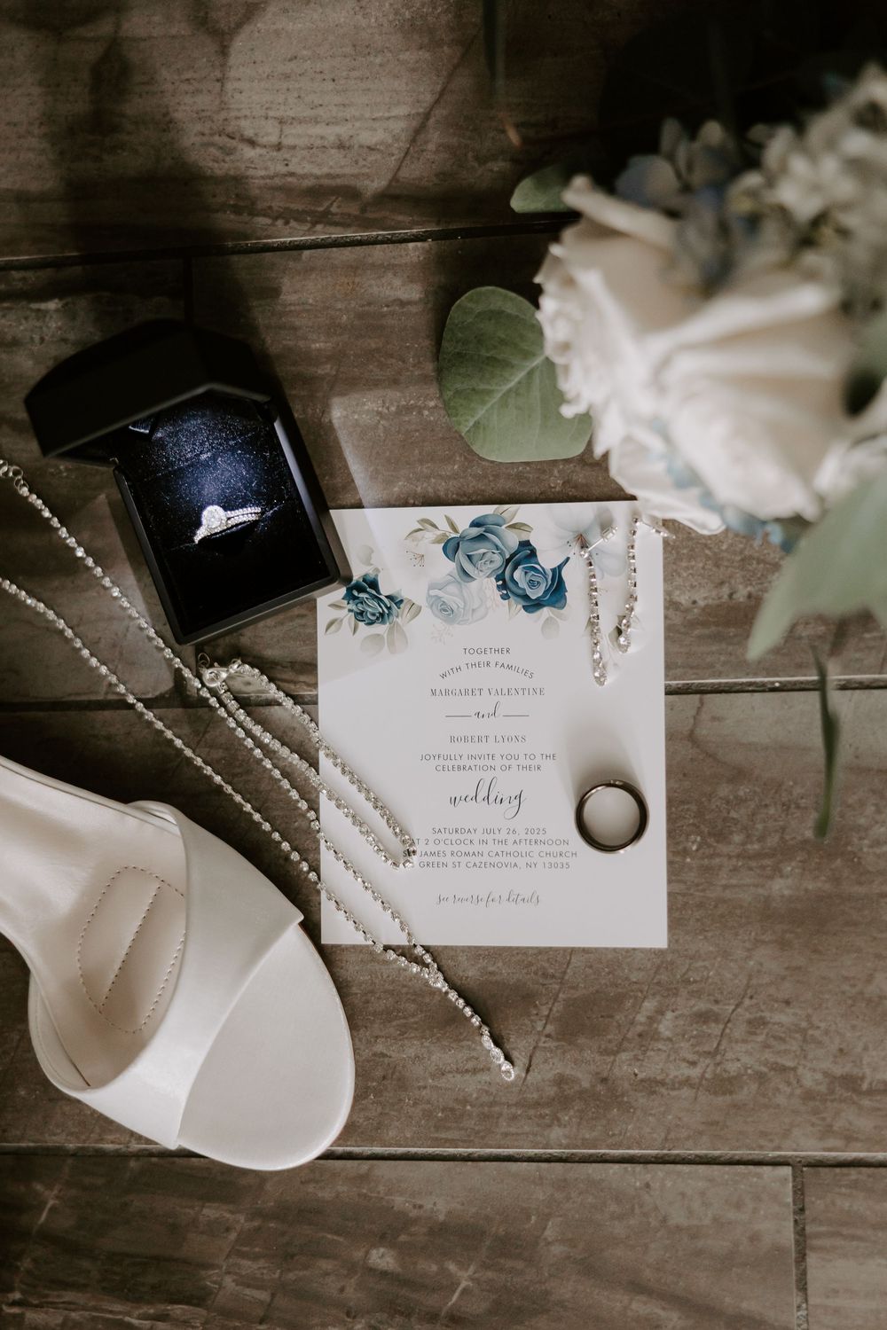 Wedding flat lay featuring invitation, diamond ring, pearl necklace, white heels, and floral bouquet.