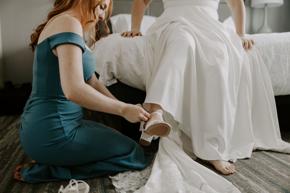 Bridesmaid in teal gown helping bride put on wedding shoes while seated on bed in bridal suite.