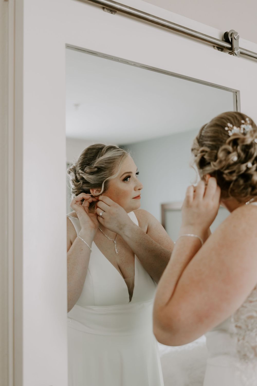 Bride in white wedding gown putting on earrings while looking in mirror during wedding preparations.