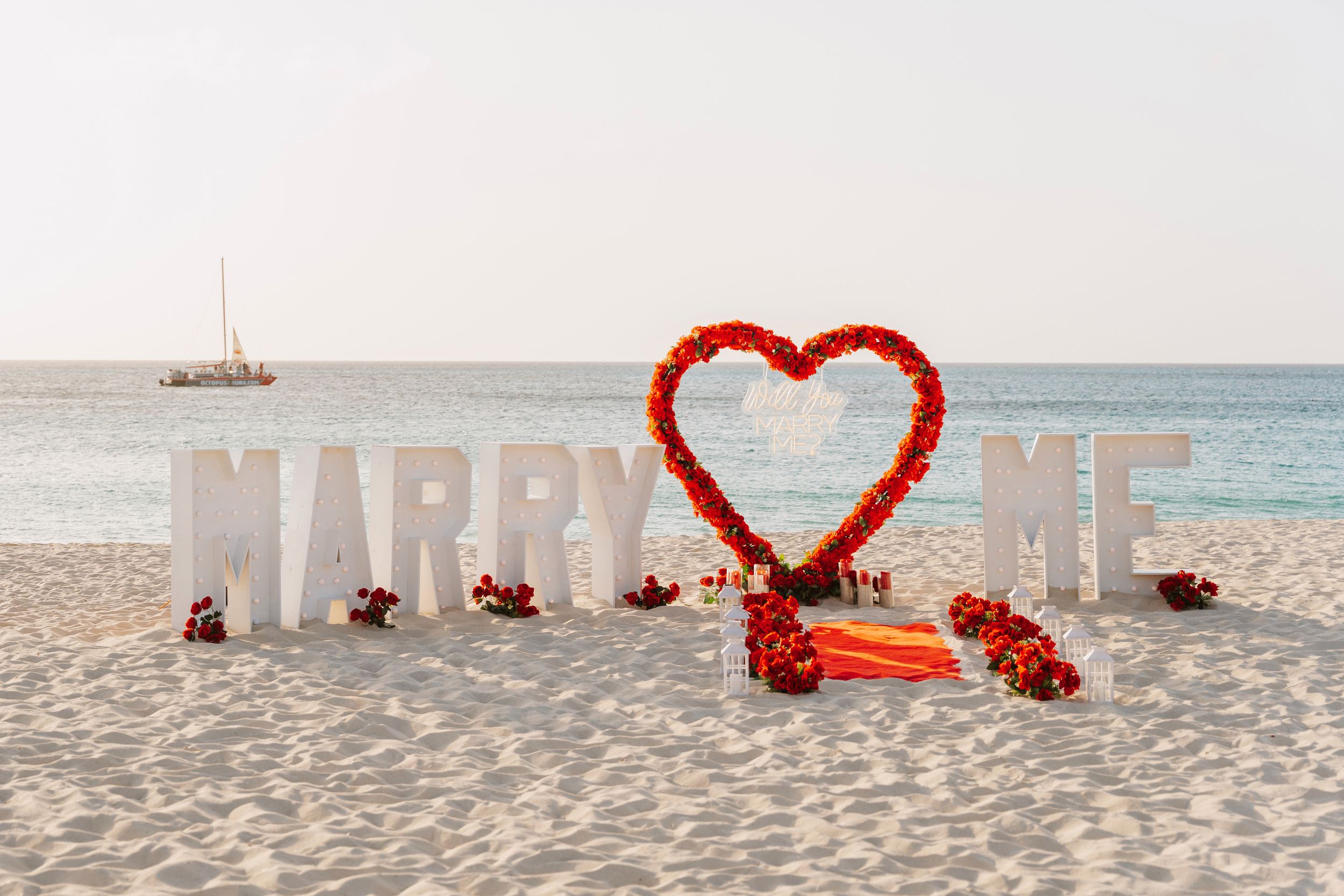 Couple holding hands at sunset beach proposal setup with floral arch, candles, and Marry Me sign on sandy shore.