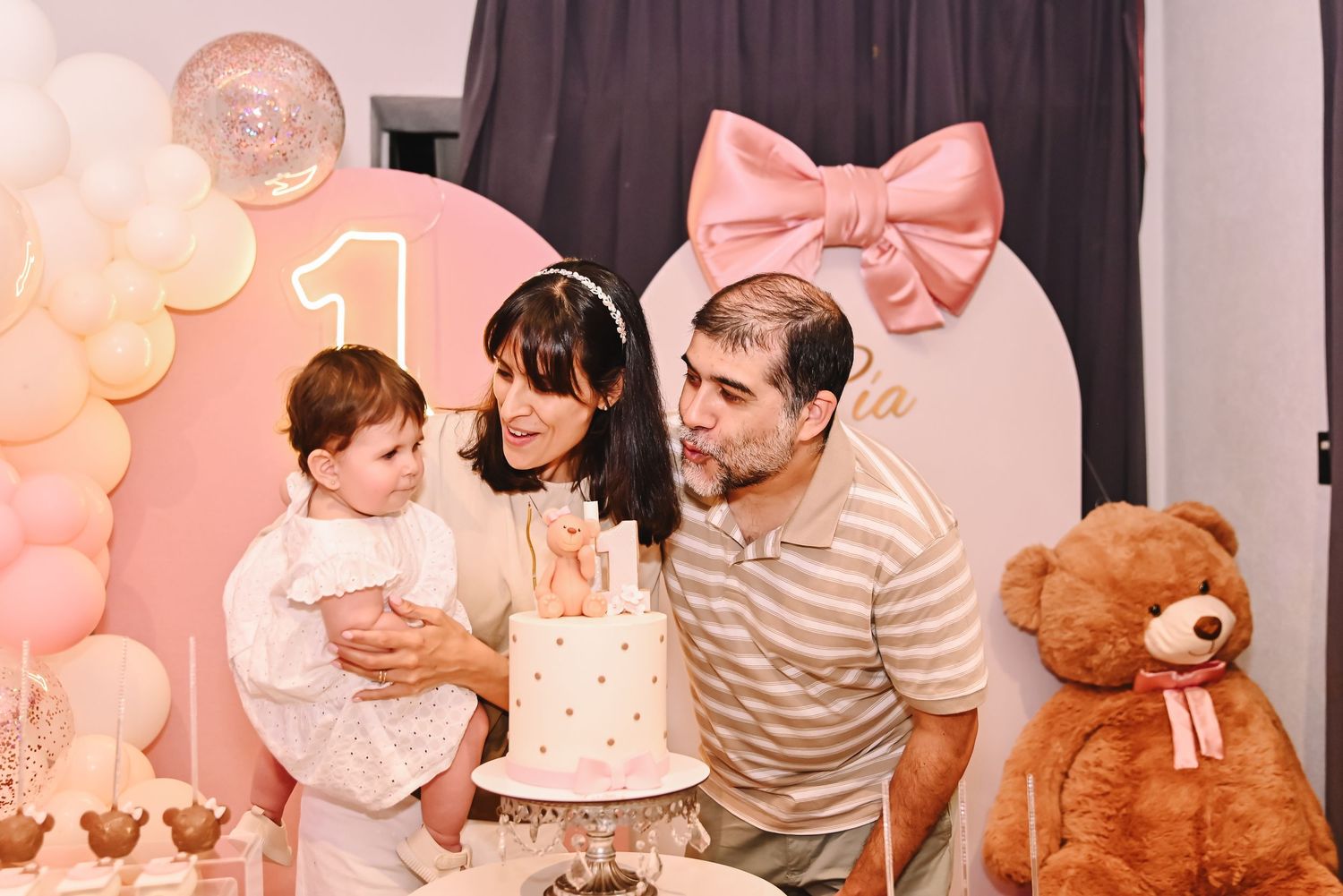 Family celebrating a baby’s first birthday party with a decorated cake table and colorful balloon arch backdrop.