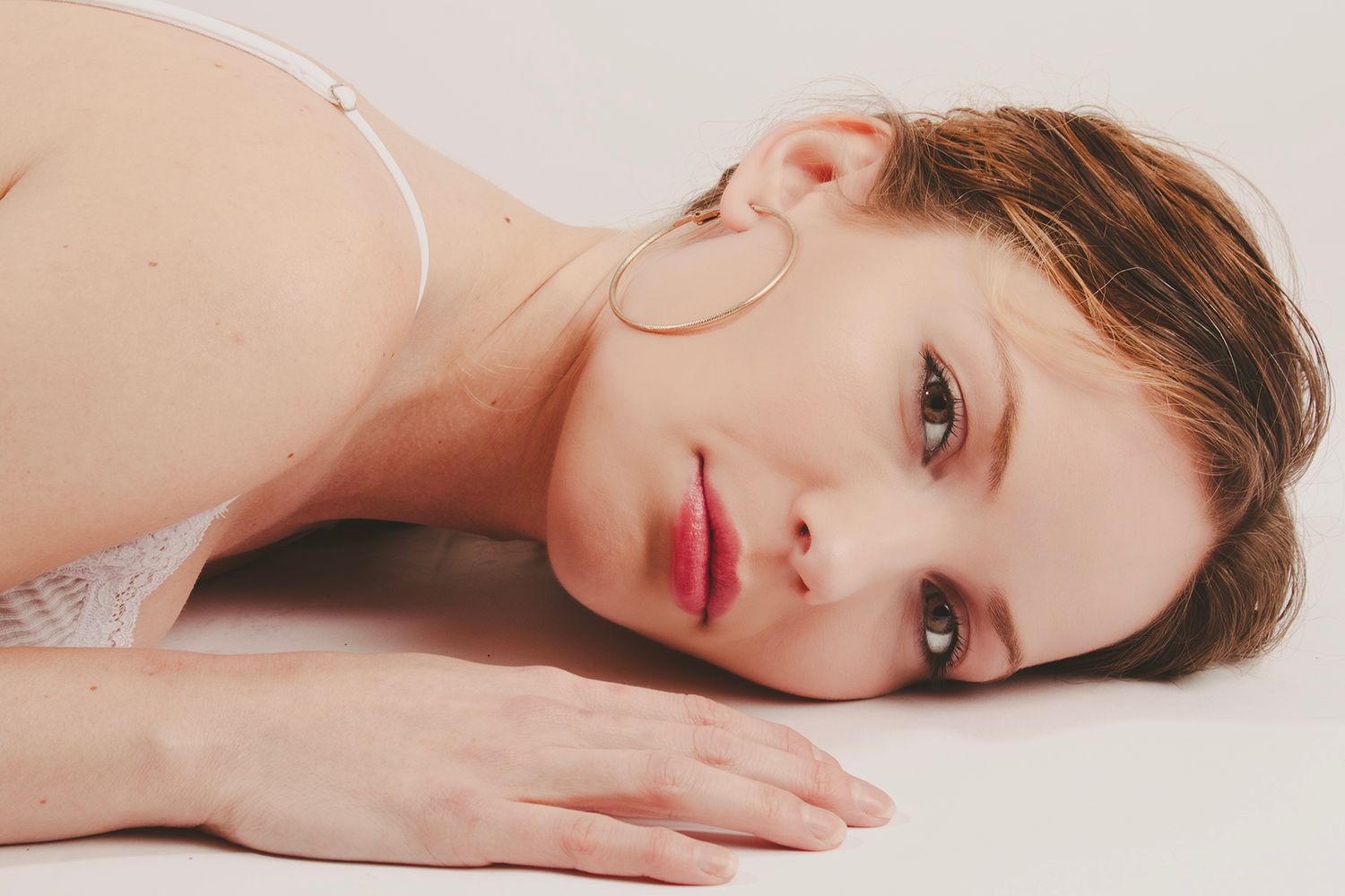Young woman with hoop earrings lies down gazing softly at camera on white background.