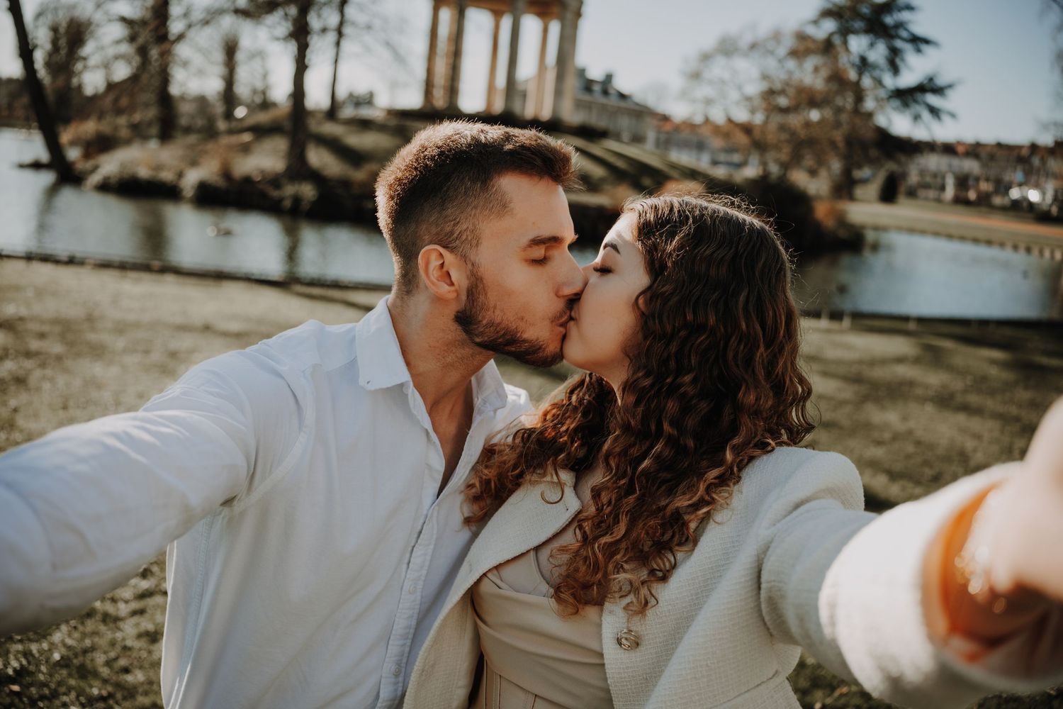 Couple sharing a romantic kiss while taking a selfie outdoors near a scenic lake and classical columns.
