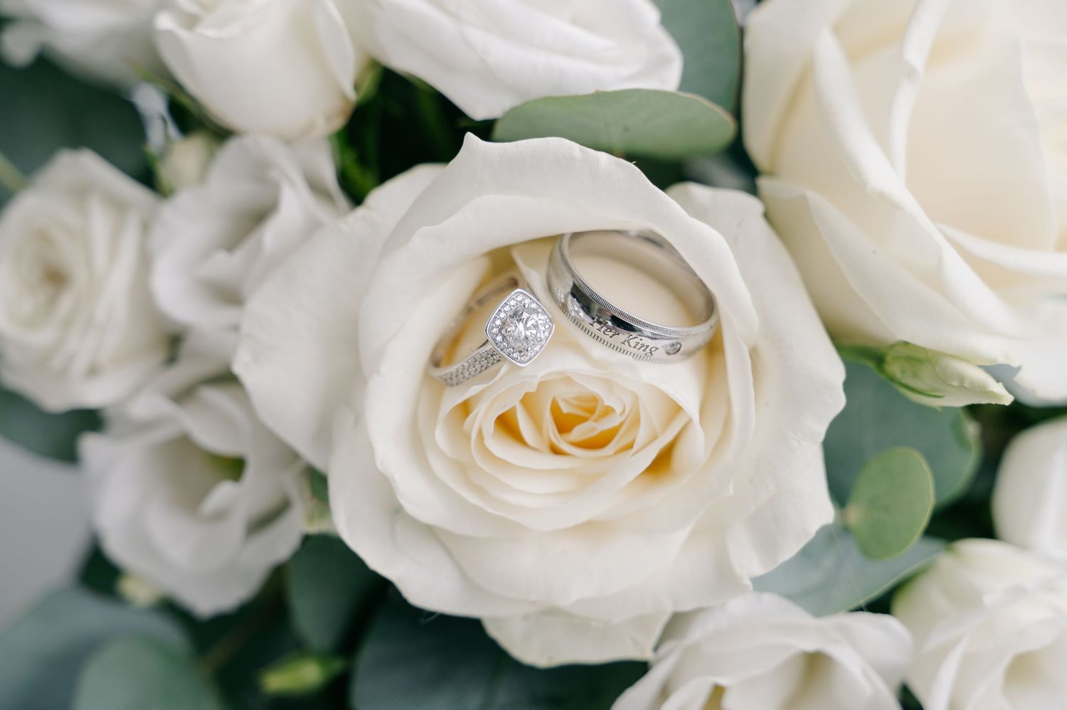 Two wedding rings nestled in a white rose bouquet, featuring a diamond engagement ring and a silver band.