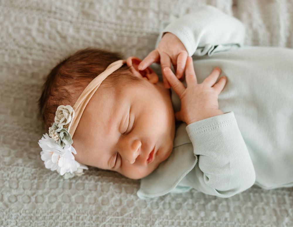Peaceful newborn baby girl sleeping in mint green outfit with floral headband on white knit blanket.