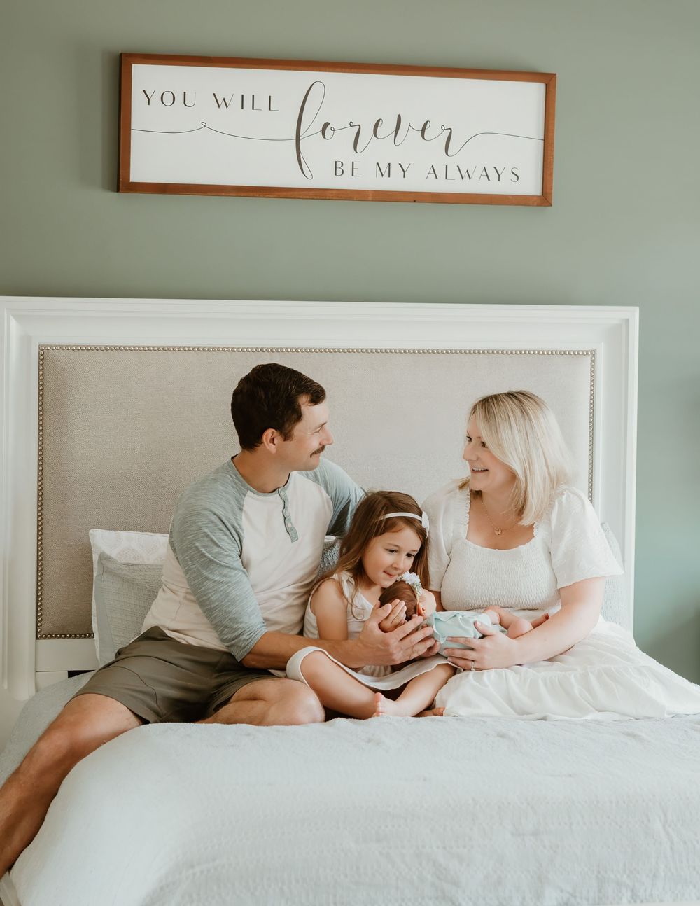 Family of four sitting on a bed beneath a Forever Be My Always sign, parents smiling at young daughter holding newborn.