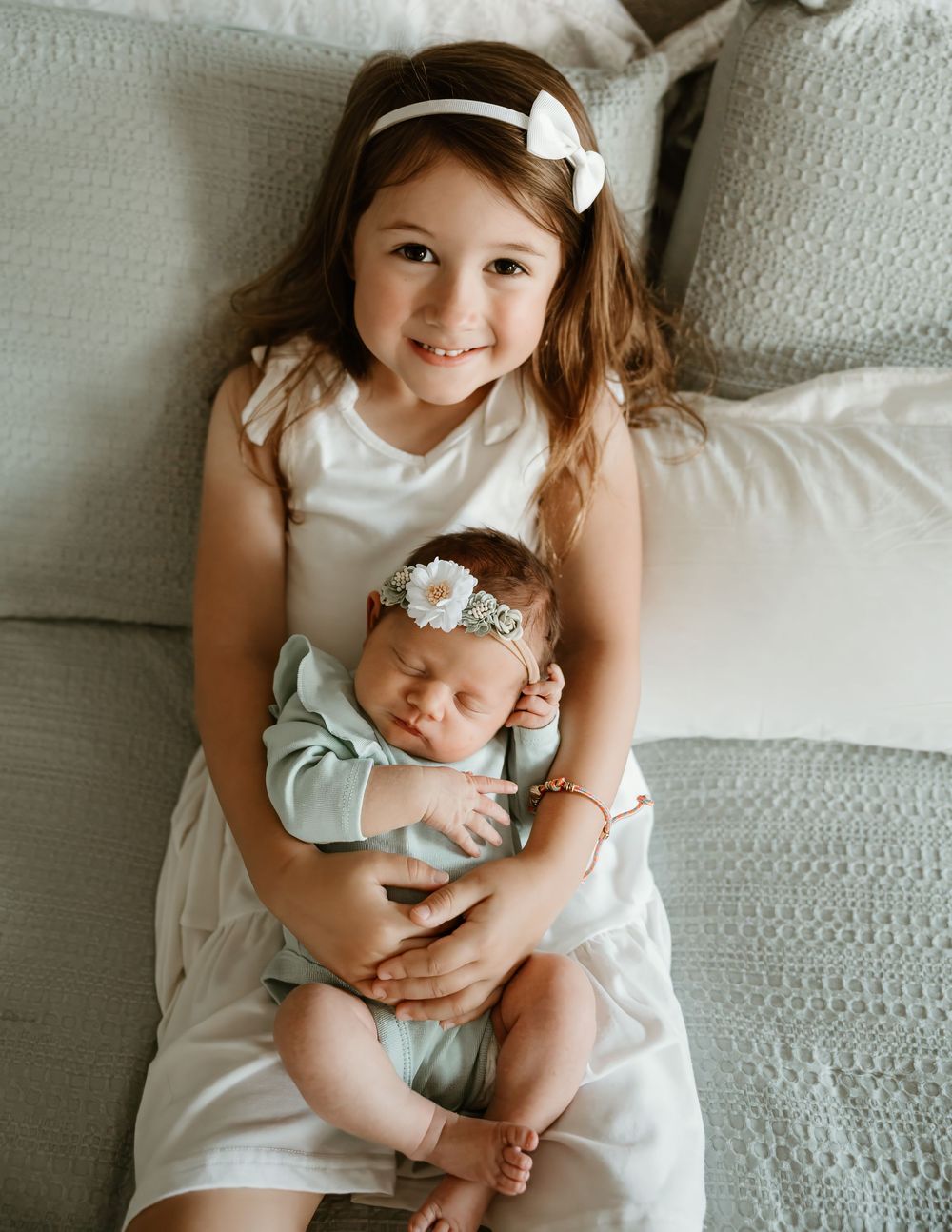Young girl in white dress smiles holding sleeping newborn baby in mint outfit on a cozy gray couch.