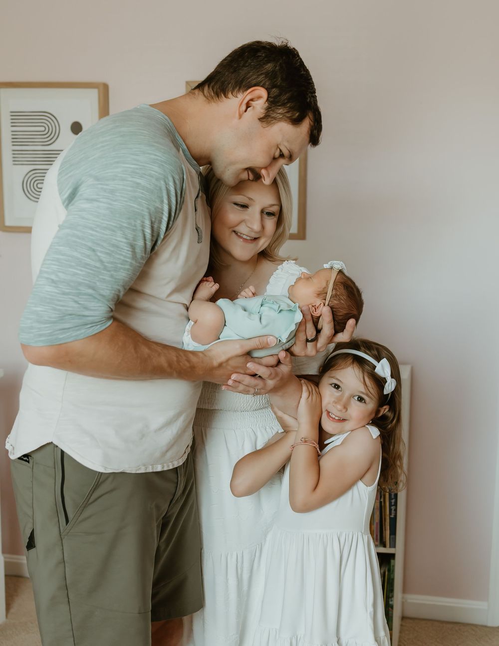 A happy family of four with a dad, mom, newborn baby, and young daughter in white dress posing together indoors.