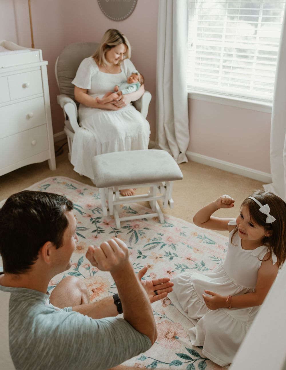 Family in a nursery, mom holding newborn in rocking chair while dad and daughter play on floral rug.