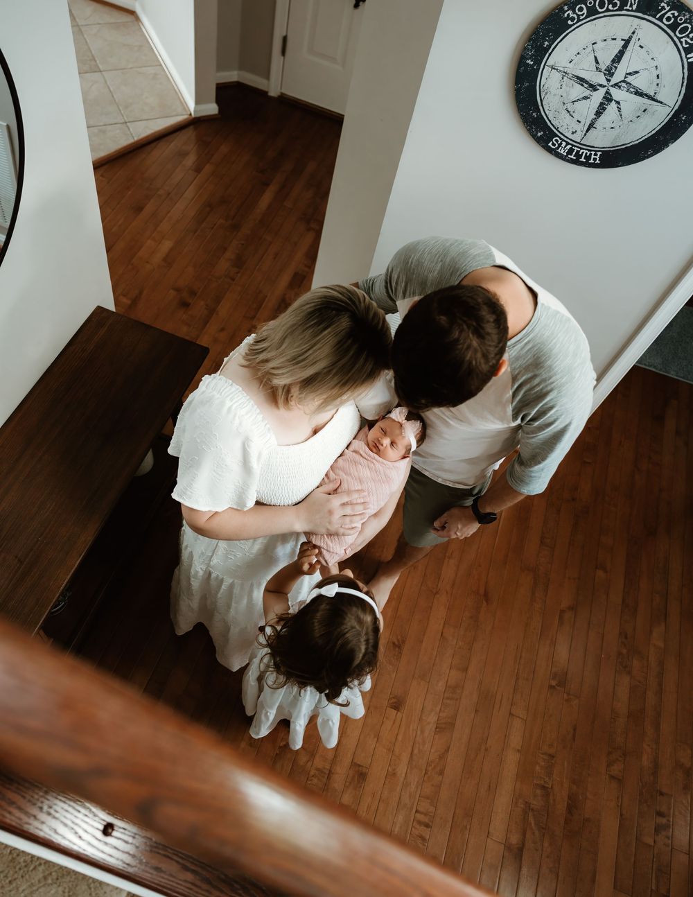 Aerial view of parents holding newborn baby with toddler daughter standing on hardwood floor.