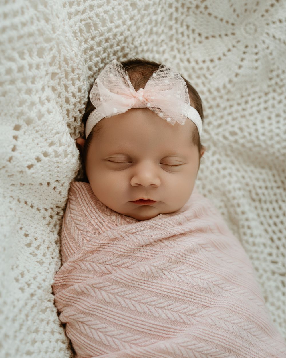 Sleeping newborn baby girl swaddled in pink wrap with bow headband on white crochet blanket.