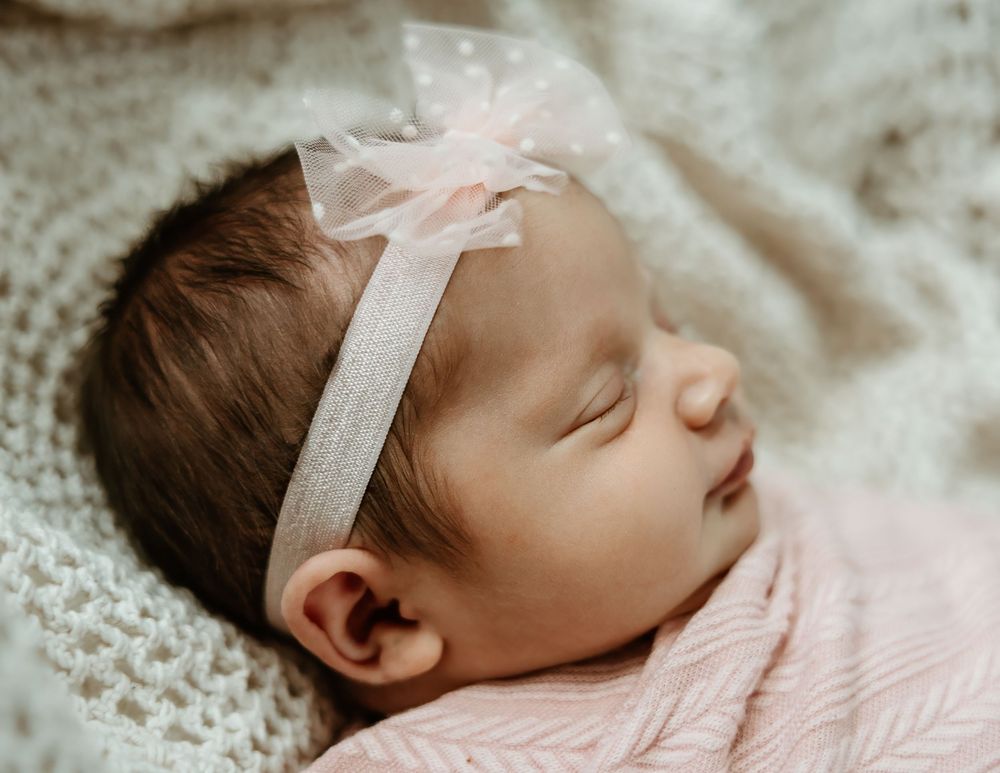 Peaceful newborn baby girl sleeping wrapped in pink swaddle with delicate blush bow headband on knit blanket.