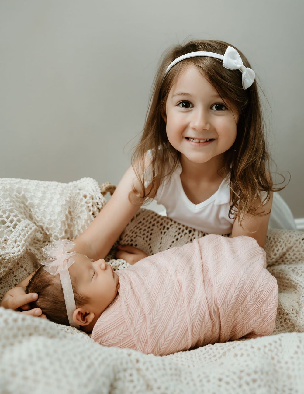 Young girl with white bow headband smiling beside sleeping swaddled newborn baby.