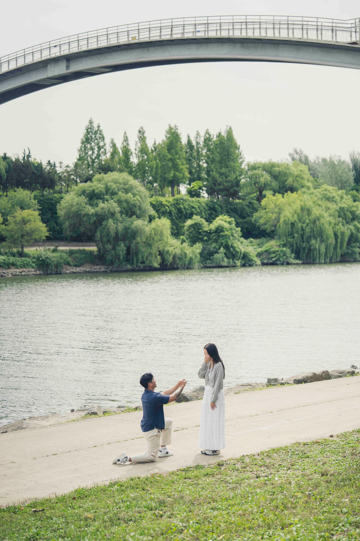Man proposes to woman by a serene river with an arched bridge and lush green trees in the background.