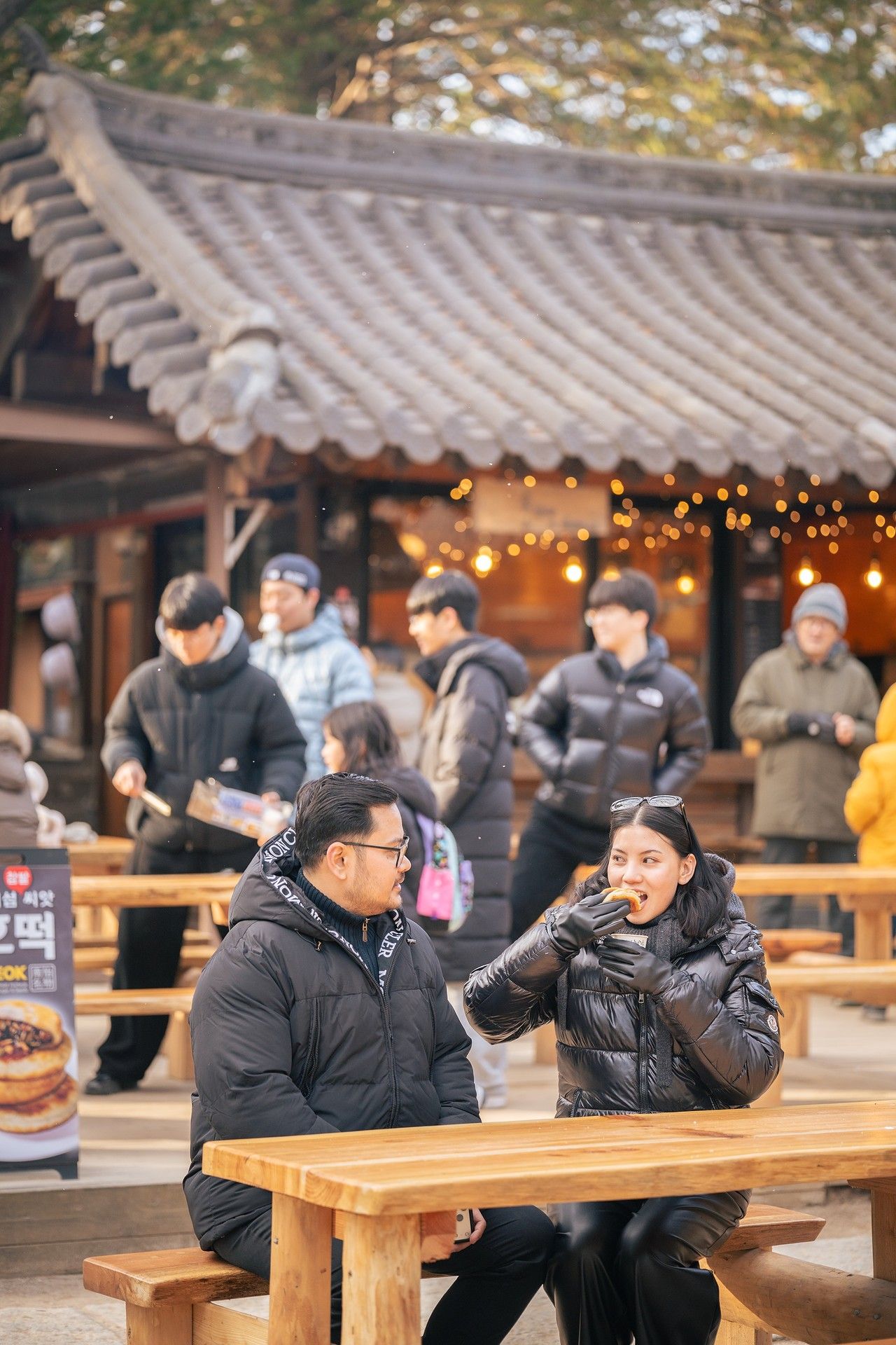 Two people in black winter jackets eating at a wooden table outside a traditional Korean building.