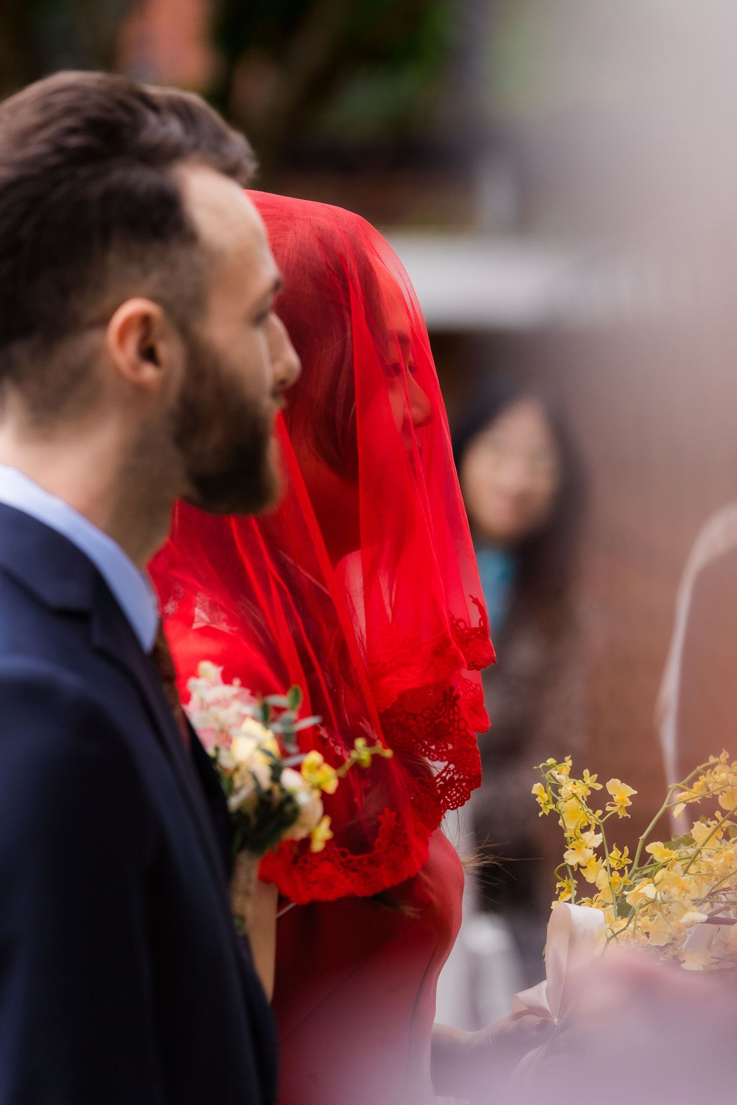 Bearded groom in navy suit stands beside bride wearing elegant red lace veil at wedding ceremony.
