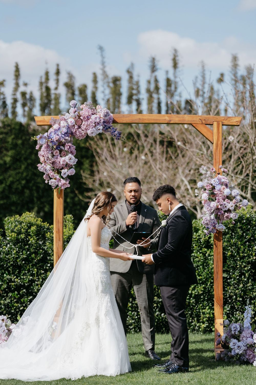 Outdoor wedding ceremony under wooden arch with purple floral arrangements, bride in white gown and groom in black suit exchanging vows.