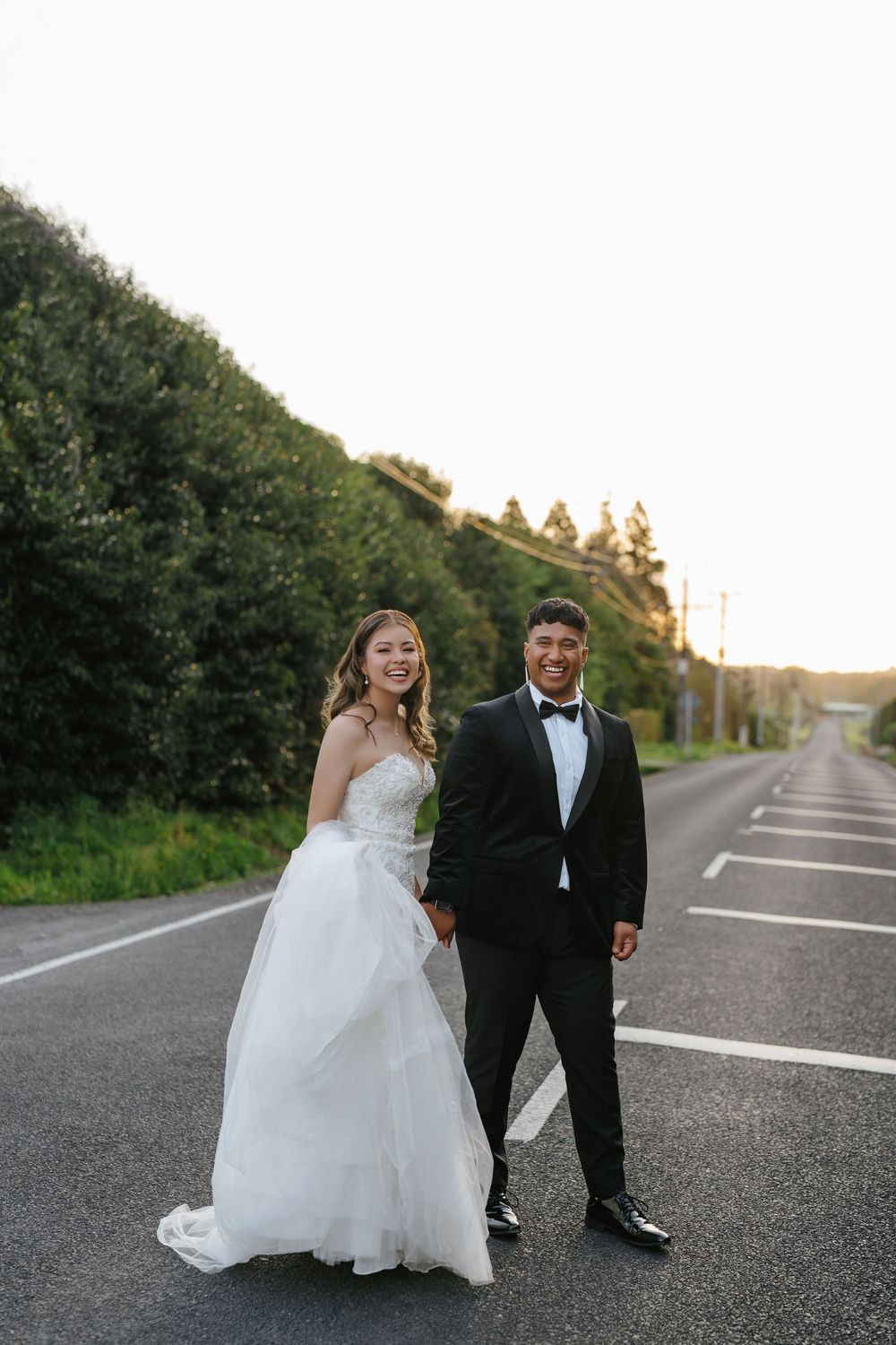 A bride in a white gown and groom in a black tuxedo hold hands on a road at sunset.