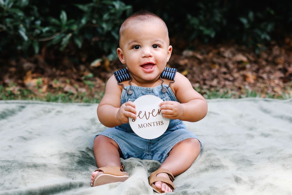 Smiling baby in denim overalls holding a seven months milestone sign, sitting outdoors on a blanket.