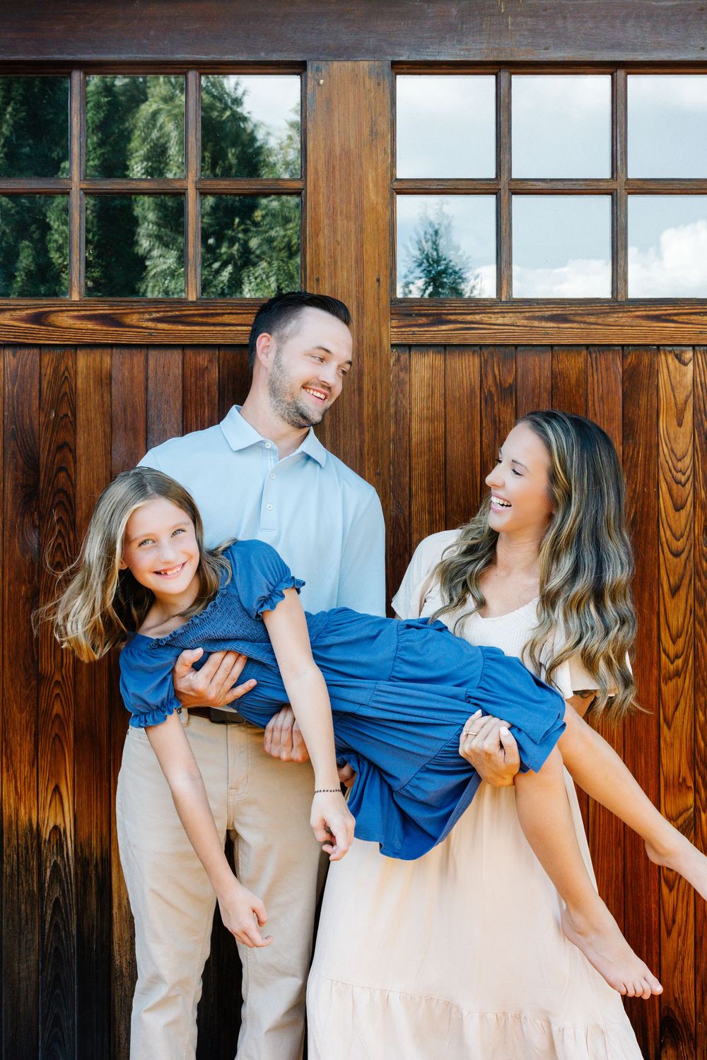 A joyful family of three poses in front of a wooden barn door, with parents lifting their smiling blonde daughter in a blue dress.