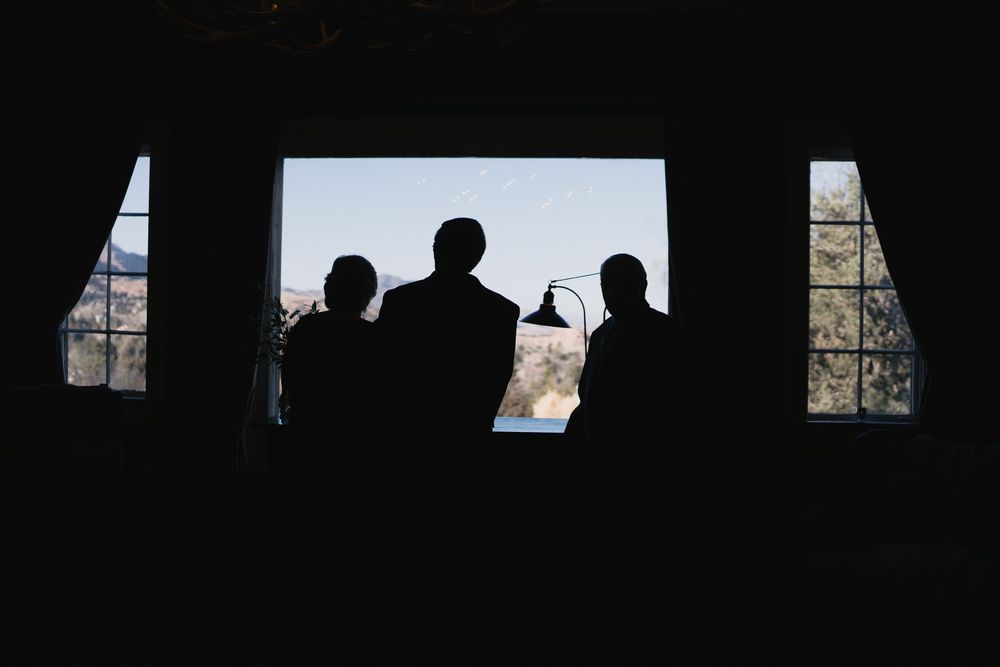 Silhouettes of three people standing before a large window with a scenic outdoor view in a dimly lit room.