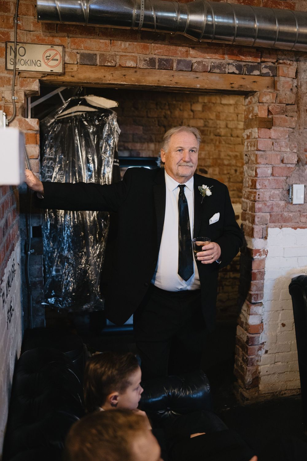 Elderly man in black suit with boutonniere holds drink, standing by brick wall at formal event.
