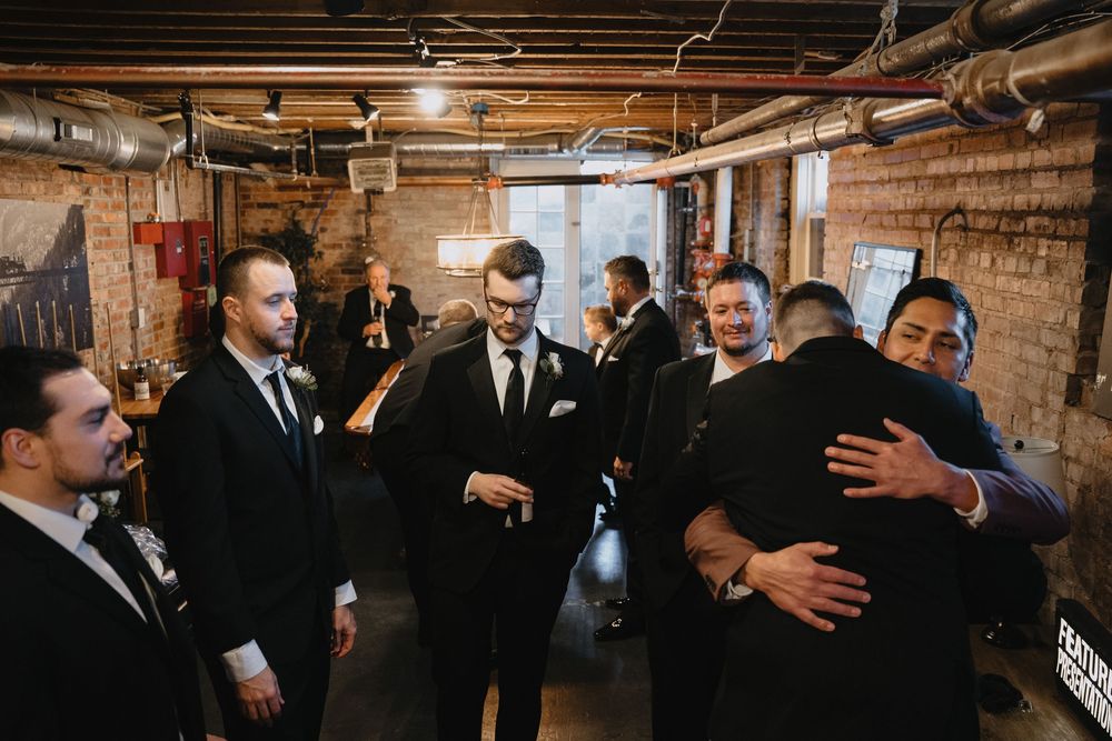 Groomsmen in black suits gather in a rustic brick venue, sharing hugs and laughter before a wedding ceremony.