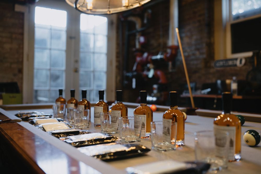 Whiskey bottles and tasting glasses lined up on a bar counter in a rustic brick-walled distillery tasting room.