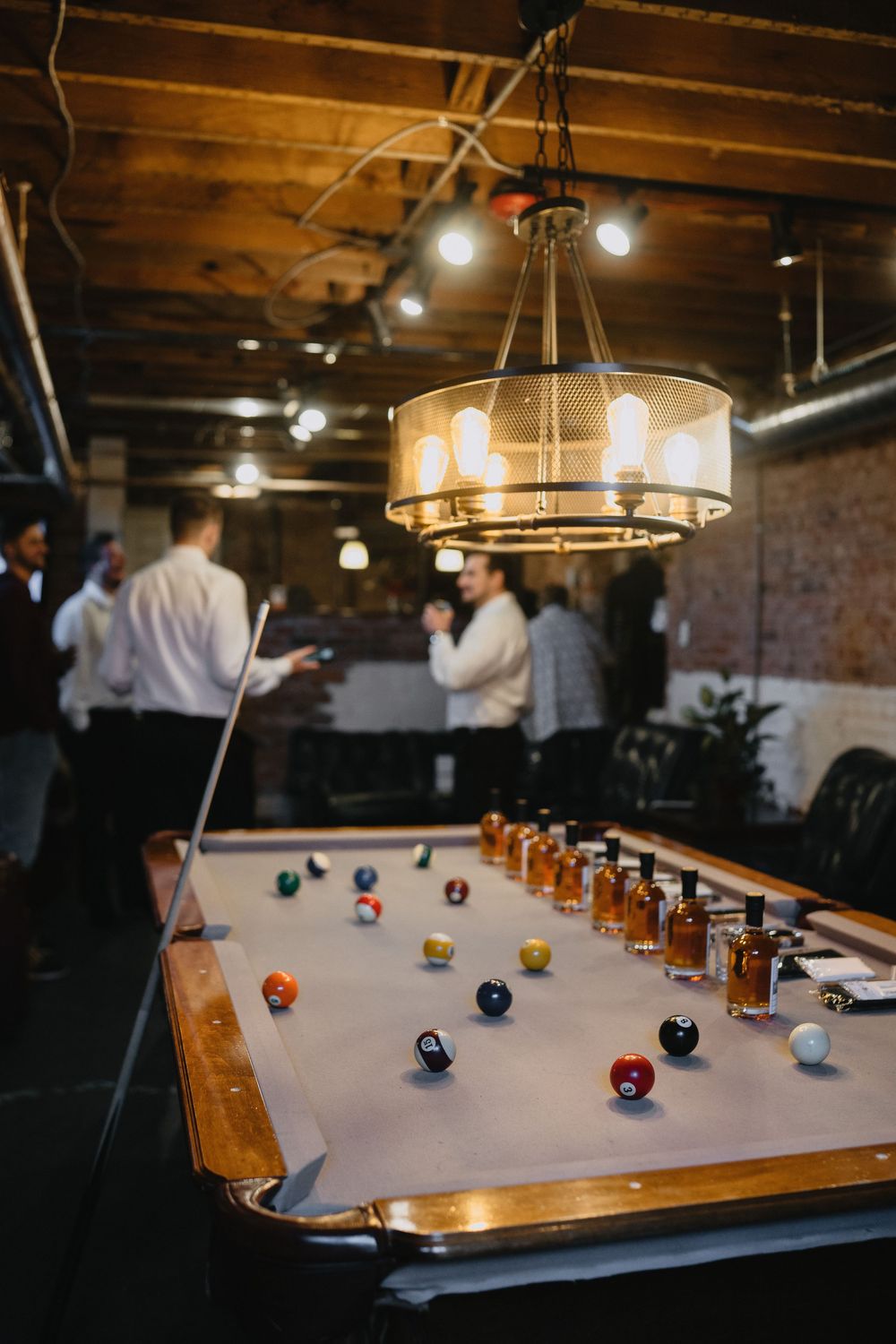Billiard room with pool table, colorful balls, whiskey shots, pendant light, and people socializing in background.
