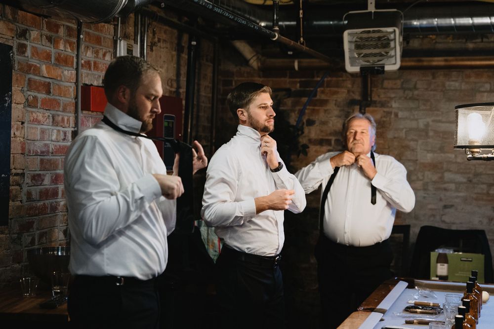 Three men in white dress shirts adjusting their ties and cufflinks in a rustic brick-walled room before a formal event.