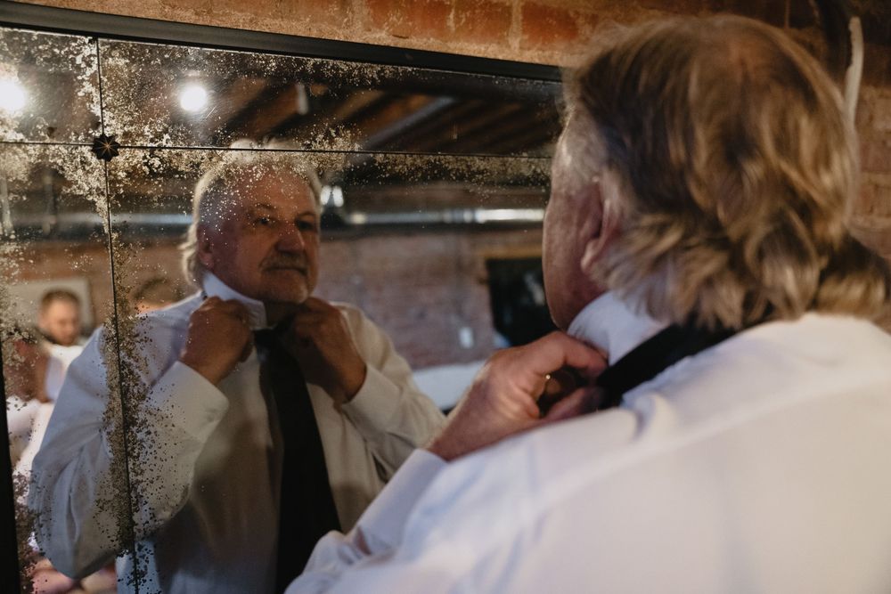 Older man adjusting his tie while looking in an antique mirror before a wedding.
