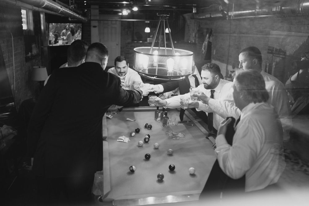 Black and white photo of men gathered around a table playing a game in a dimly lit room with a chandelier.