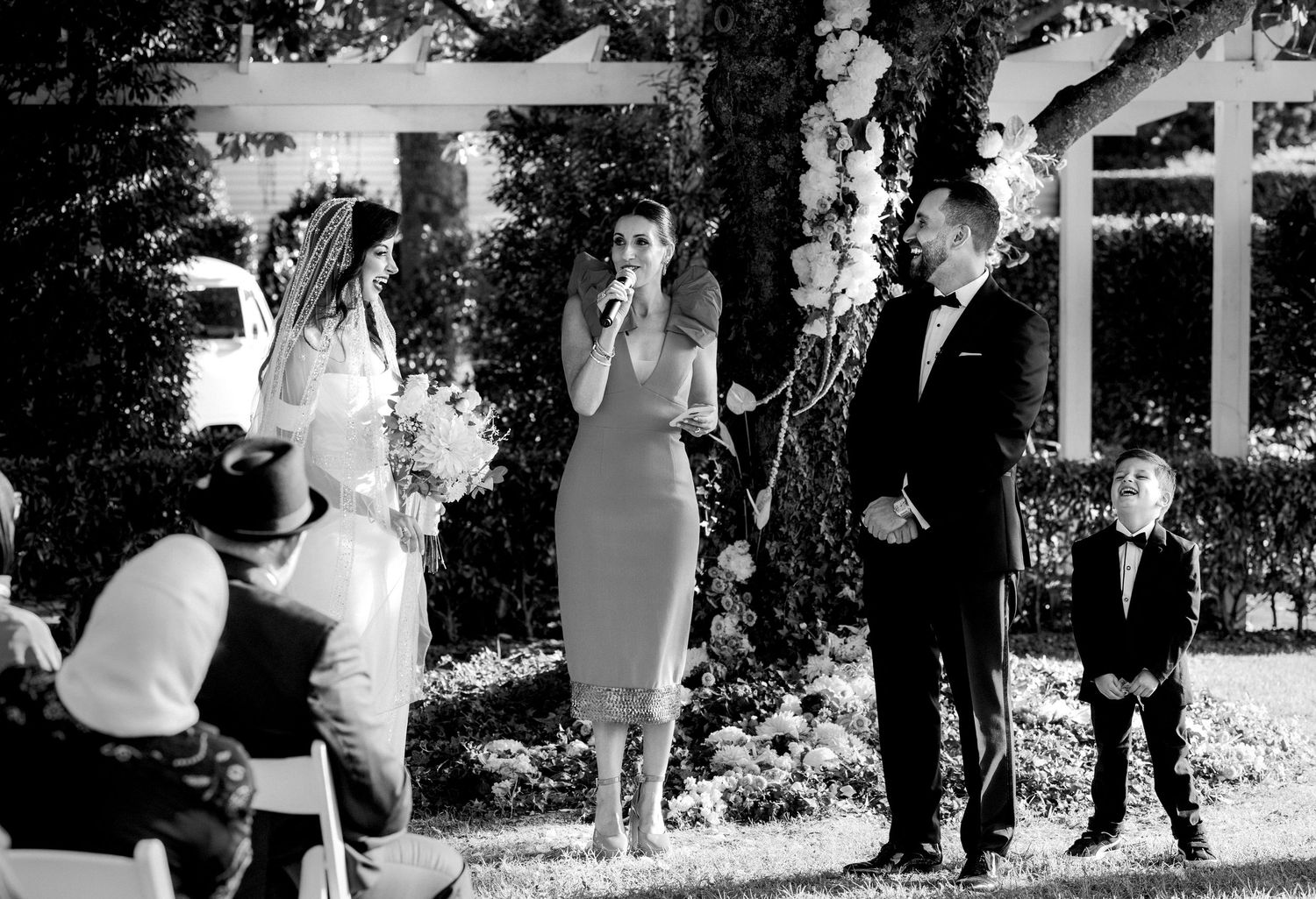 Black and white outdoor wedding ceremony with bride, officiant speaking into mic, groom, and young boy in suit.