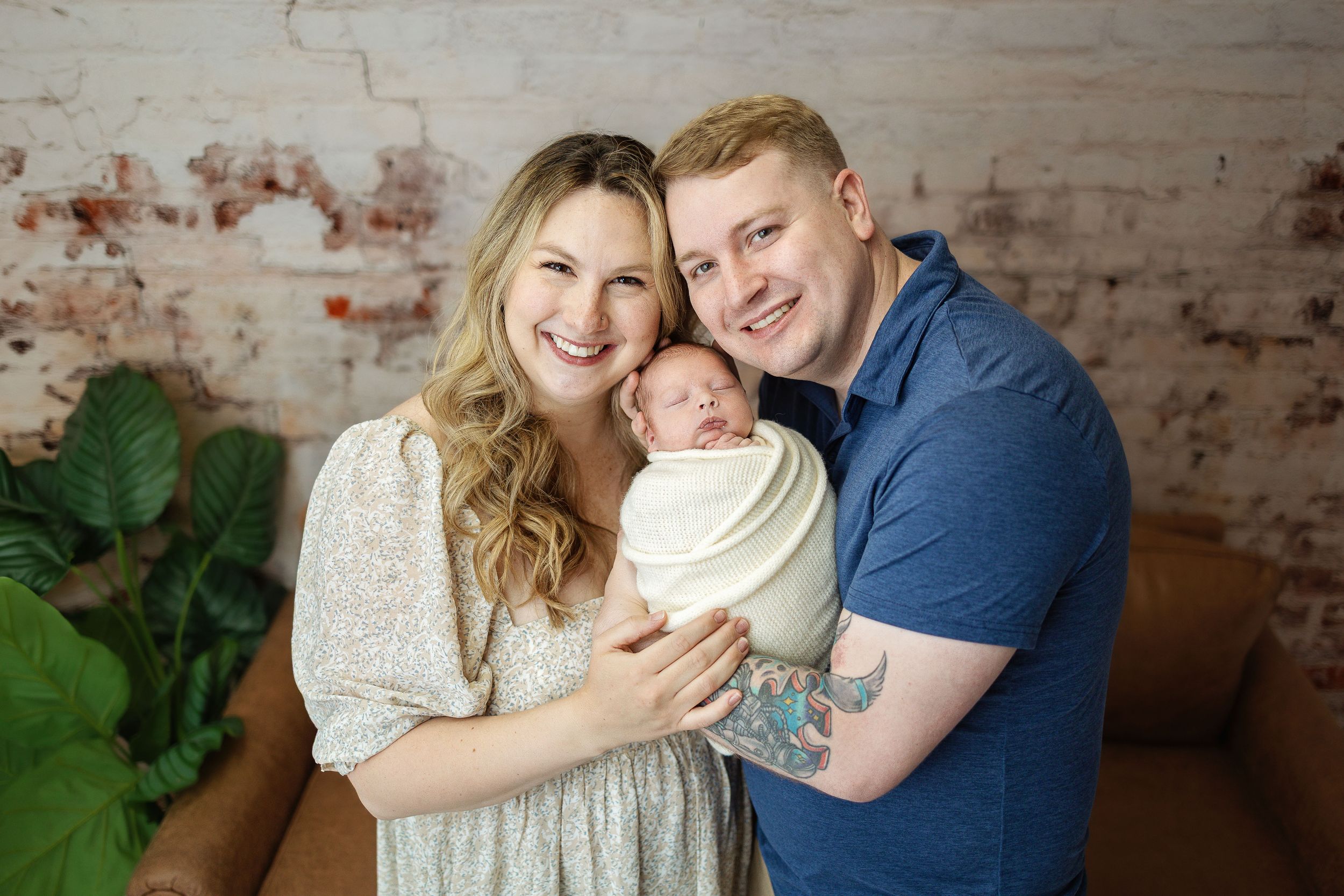 Smiling couple holding swaddled newborn baby in front of rustic brick wall in professional studio portrait.