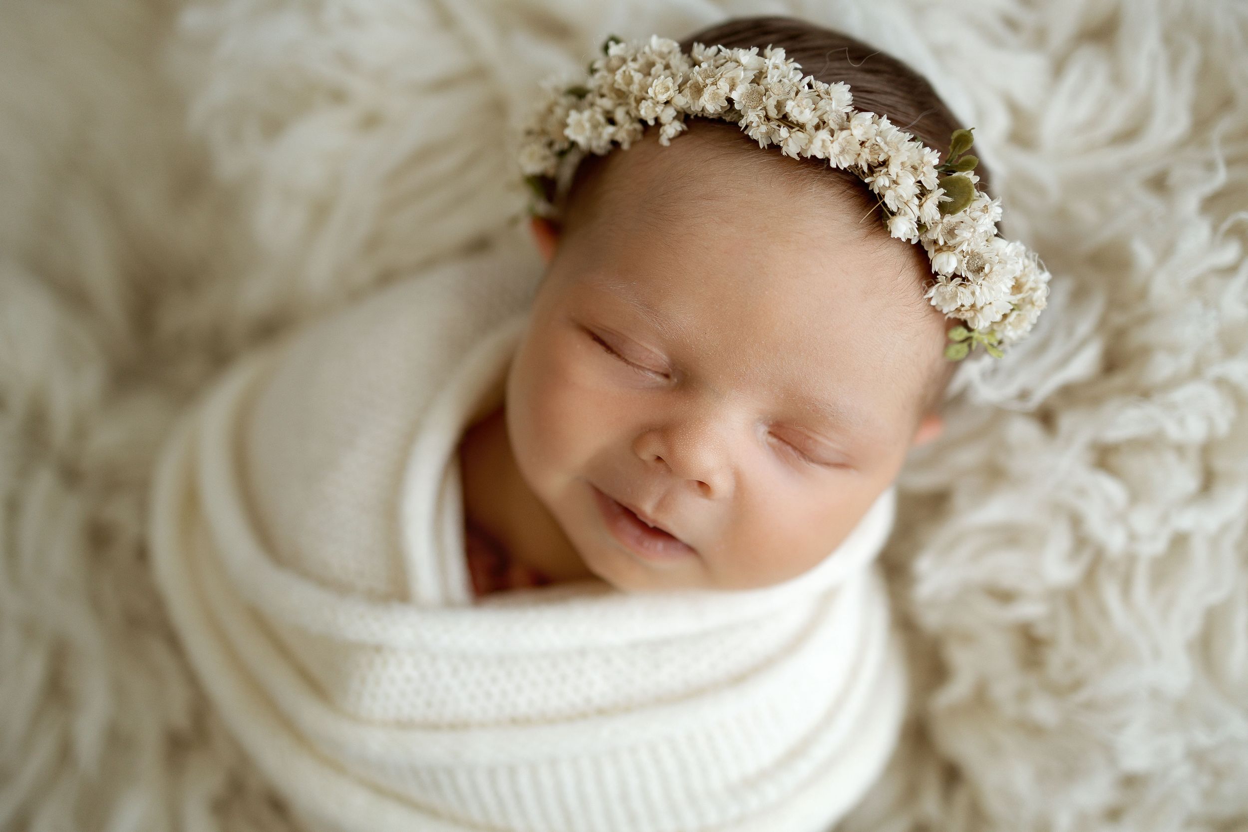 Sleeping newborn baby swaddled in white wrap wearing a delicate floral crown on a fluffy cream blanket.