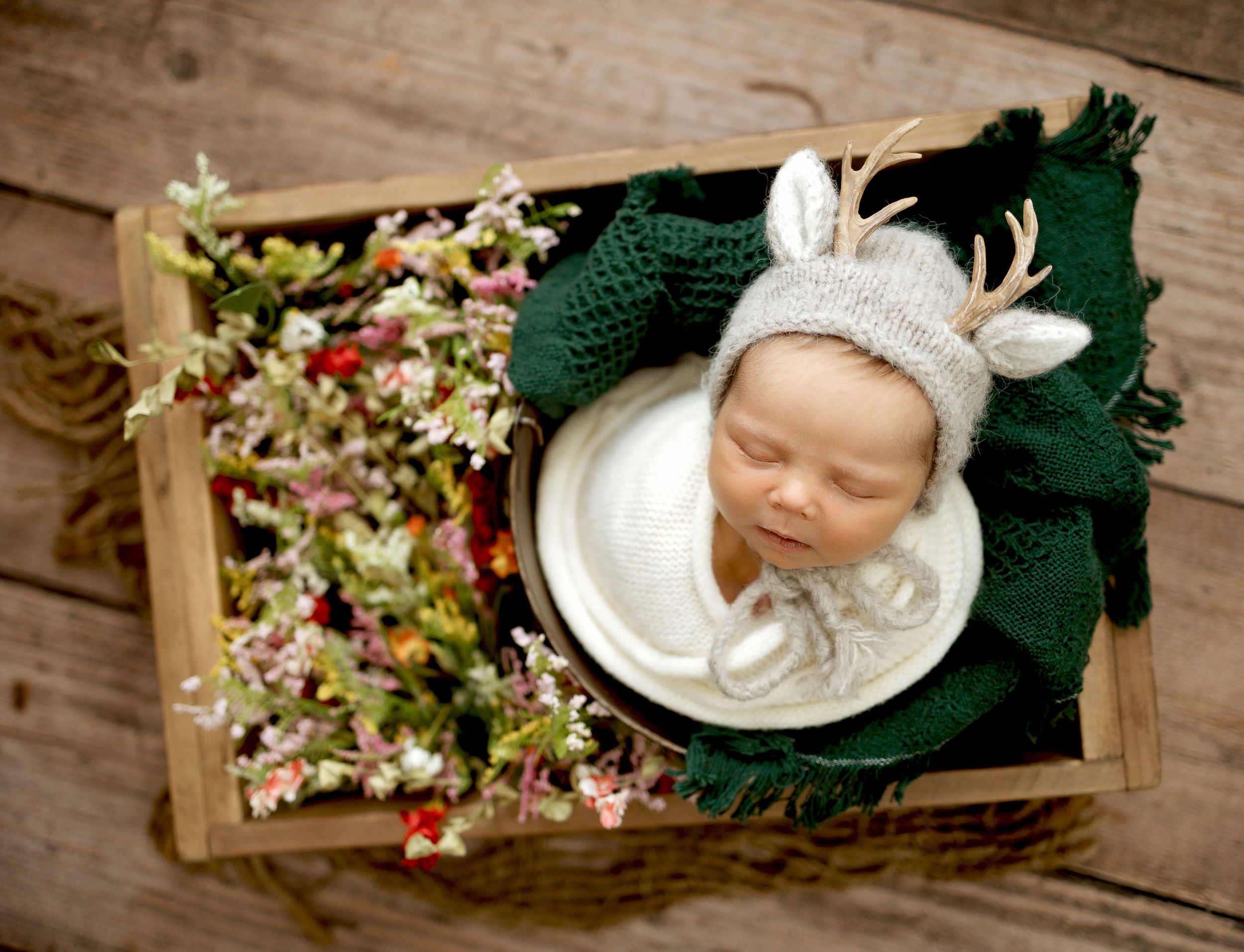 Newborn baby sleeping in a white bowl wearing a knit reindeer hat, surrounded by flowers in a rustic wooden crate.