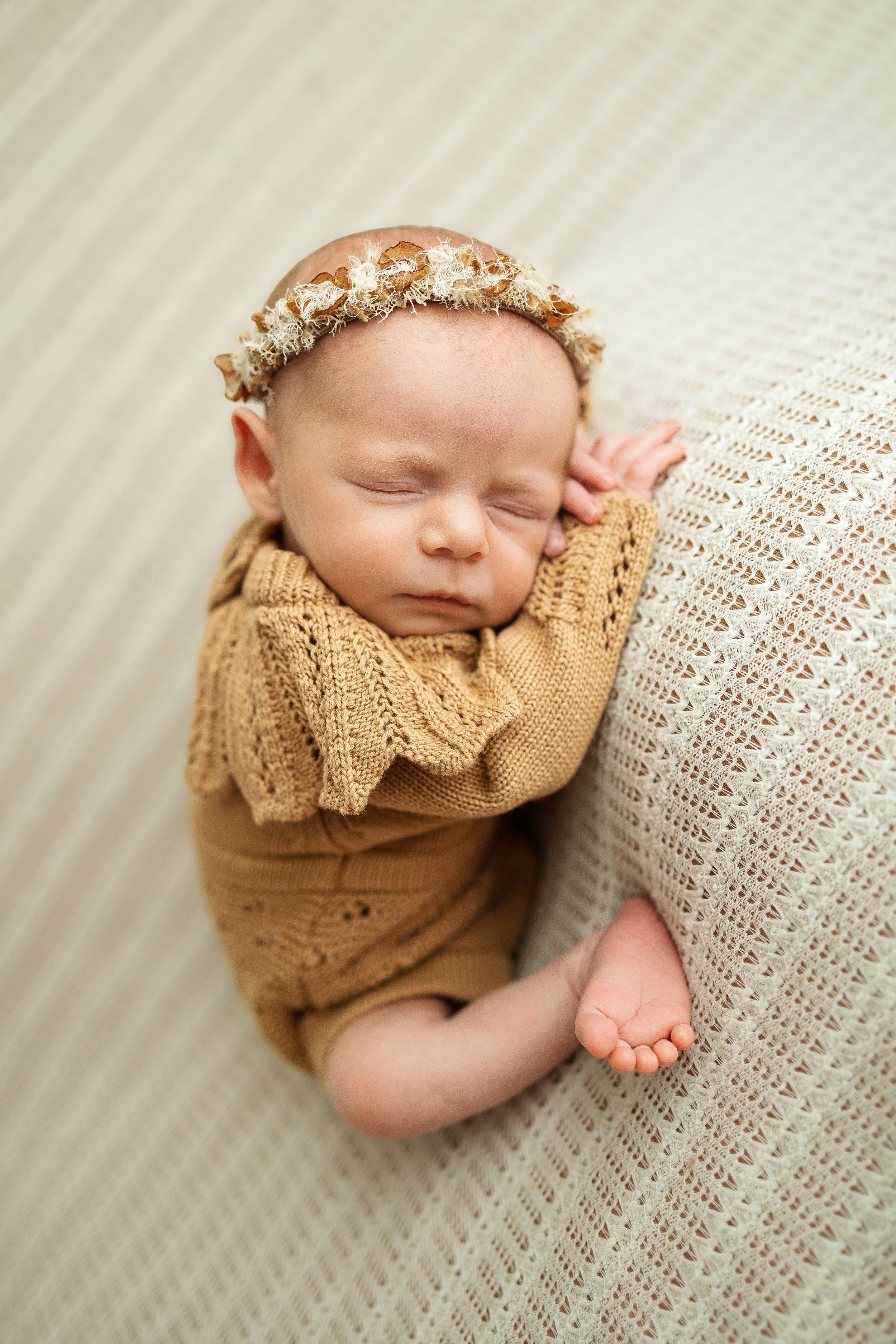Sleeping newborn baby wrapped in a golden knit blanket wearing a delicate floral headband on a cream textured surface.