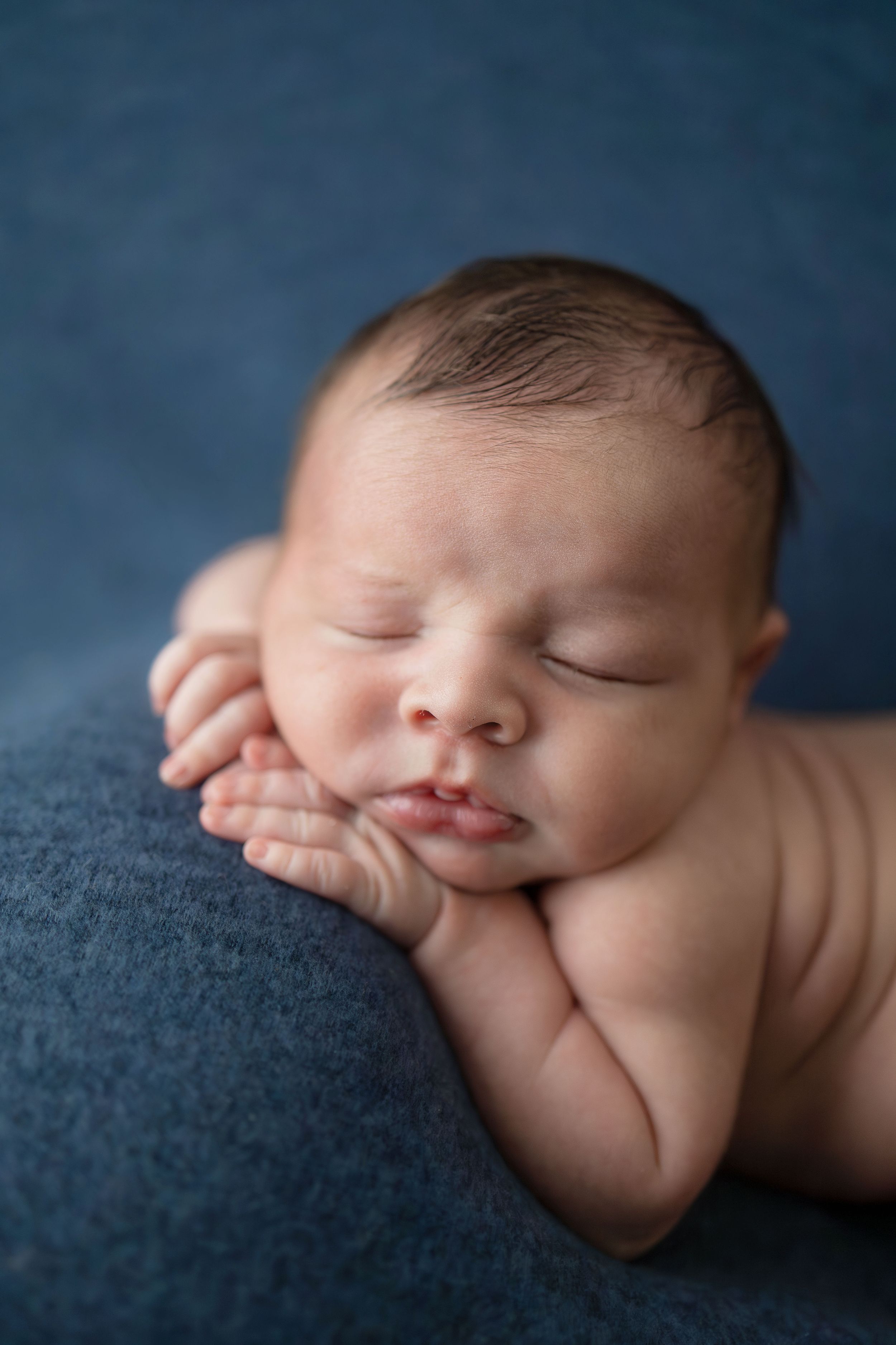 Peaceful newborn baby sleeping on blue fabric with hands tucked under chin in classic posed newborn photography.